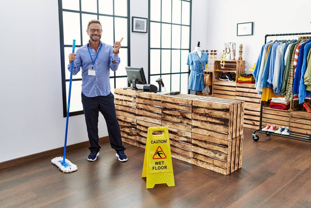 Mann mittleren Alters, der in einer Boutique arbeitet und den Boden putzt, lächelt glücklich und zeigt mit der Hand und dem Finger zur Seite  - Foto, Bild
