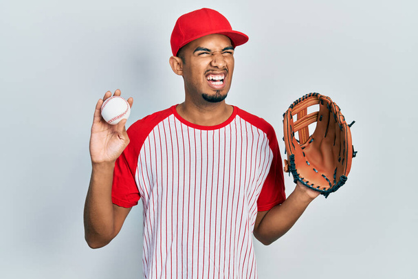 Joven hombre afroamericano vistiendo uniforme de béisbol sosteniendo guante y pelota enojado y loco gritando frustrado y furioso, gritando con ira. rabia y concepto agresivo.  - Foto, Imagen