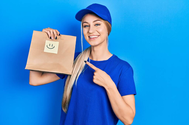 Young caucasian woman holding take away paper bag with happy face reminder smiling happy pointing with hand and finger  - Fotografie, Obrázek