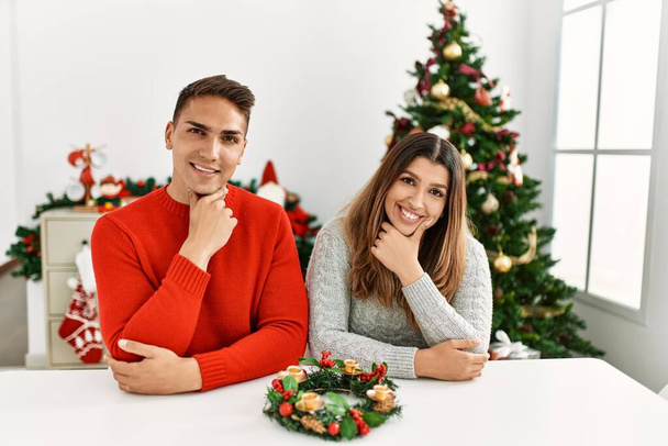 Fiatal spanyol pár ül az asztalnál karácsonykor, magabiztosan nézik a kamerát, keresztbe tett kézzel mosolyognak, és felemelt kézzel állnak. pozitív gondolkodás.  - Fotó, kép