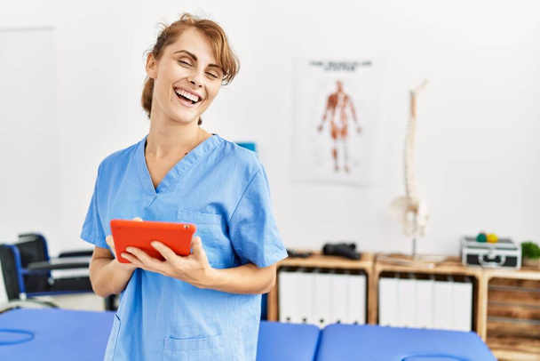Νεαρή καυκάσια φυσιοθεραπεύτρια χαμογελά ευτυχισμένη χρησιμοποιώντας touchpad στην κλινική. - Φωτογραφία, εικόνα