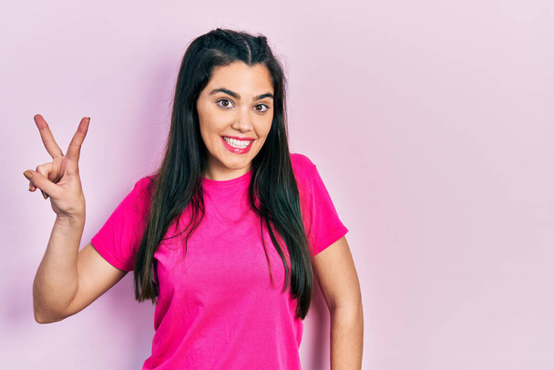 Young hispanic girl wearing casual pink t shirt smiling looking to the camera showing fingers doing victory sign. number two.  - Photo, Image