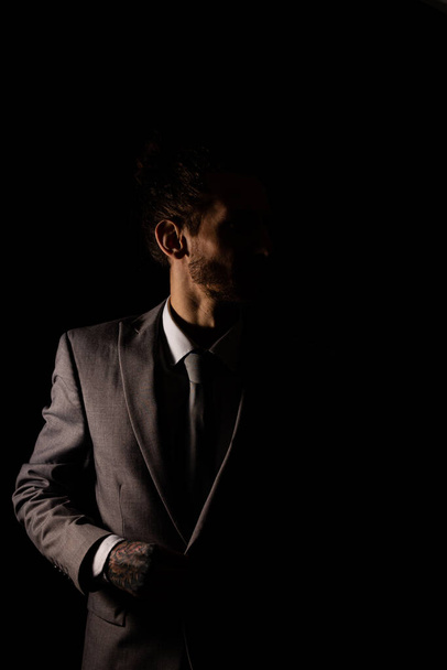 Un joven hombre de negocios con un elegante traje gris mirando a la cámara con una luz malhumorada que proyecta a la mitad de él en la sombra - Foto, Imagen