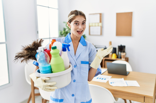 Young blonde woman wearing cleaner uniform holding cleaning products pointing aside with hands open palms showing copy space, presenting advertisement smiling excited happy  - Φωτογραφία, εικόνα