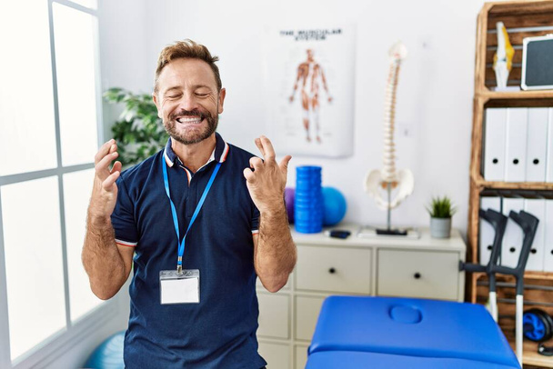 中年理学療法士の男性が痛みの回復クリニックで働いている指をジェスチャー希望と目を閉じて笑顔を越えました.幸運と迷信の概念は.  - 写真・画像