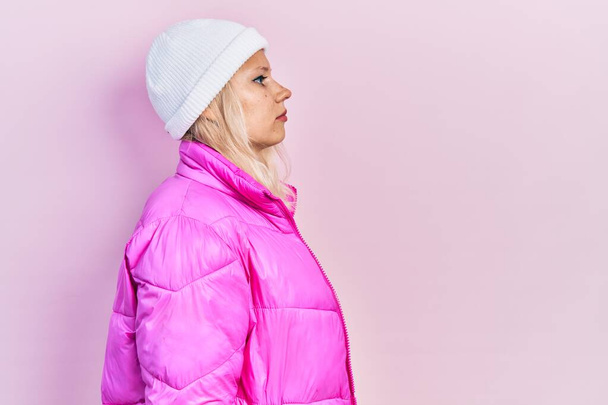 Belle femme blonde caucasienne portant un chapeau en laine et un manteau d'hiver regardant vers le côté, pose de profil relax avec un visage naturel avec un sourire confiant.  - Photo, image