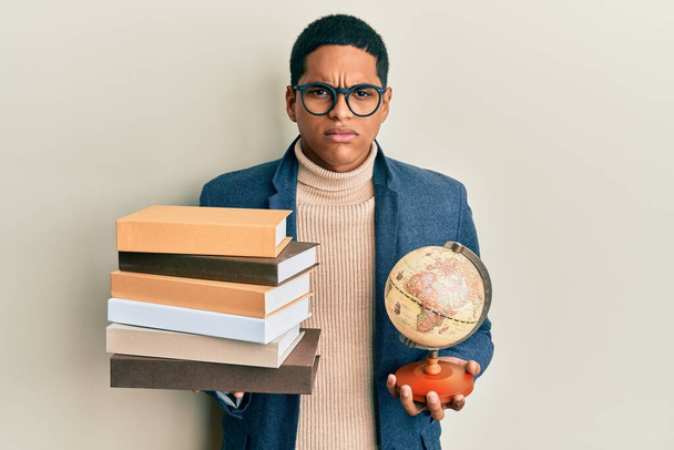 Junge gut aussehende hispanische Mann mit Vintage-Weltball und Stapel Bücher skeptisch und nervös, Stirnrunzeln aufgeregt wegen des Problems. negative person.  - Foto, Bild