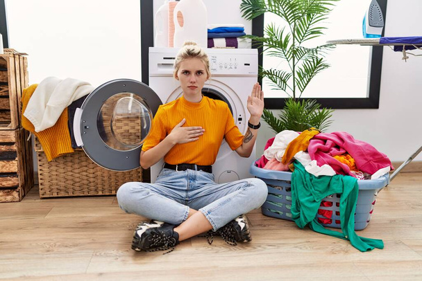 Молодая блондинка, занимающаяся стиркой, сидя у стиральной машины, ругаясь рукой на груди и открытой ладони, давая клятву верности  - Фото, изображение