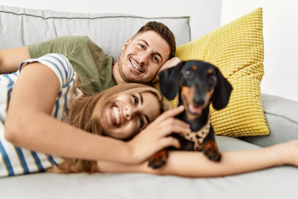 Νεαρό ζευγάρι ισπανόφωνων χαμογελαστό χαρούμενο ξαπλωμένο στον καναπέ με σκύλο στο σπίτι. - Φωτογραφία, εικόνα