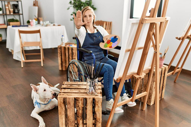 Kavkazská mladá blondýnka sedící na vozíku v ateliéru umění s otevřenou rukou, která dělá stopku s vážným a sebevědomým výrazem, obranné gesto  - Fotografie, Obrázek