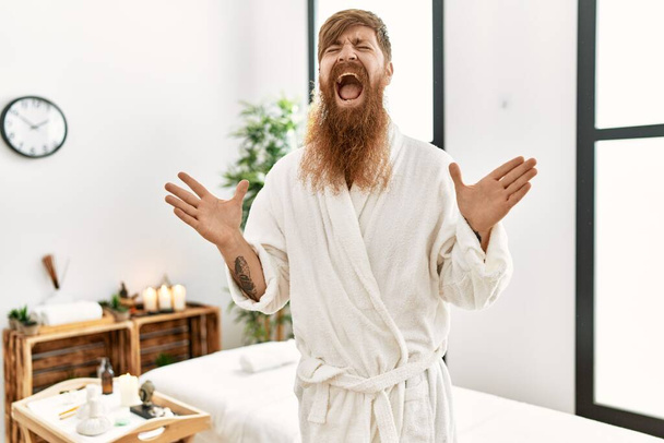 Rudy mężczyzna z długą brodą w szlafroku w spa odnowy biologicznej świętując szalony i szalony na sukces z podniesionymi ramionami i zamkniętymi oczami krzycząc podekscytowany. koncepcja zwycięzcy  - Zdjęcie, obraz