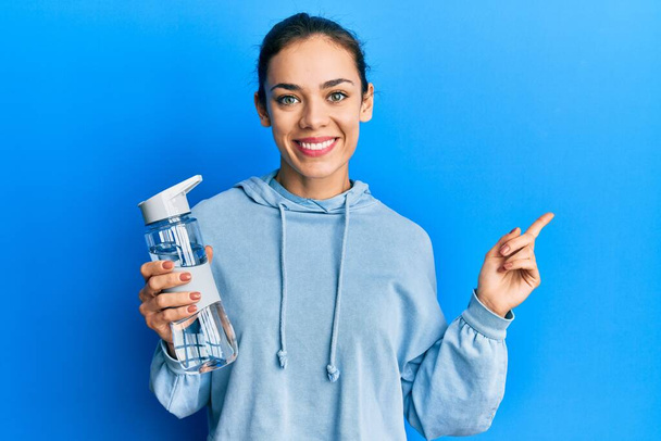 Giovane donna bionda caucasica che beve bottiglia d'acqua sorridente felice indicando con mano e dito di lato  - Foto, immagini