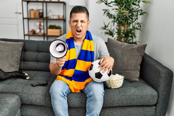 Young hispanic hooligan man supporting soccer team sitting on the sofa at home. - Photo, Image