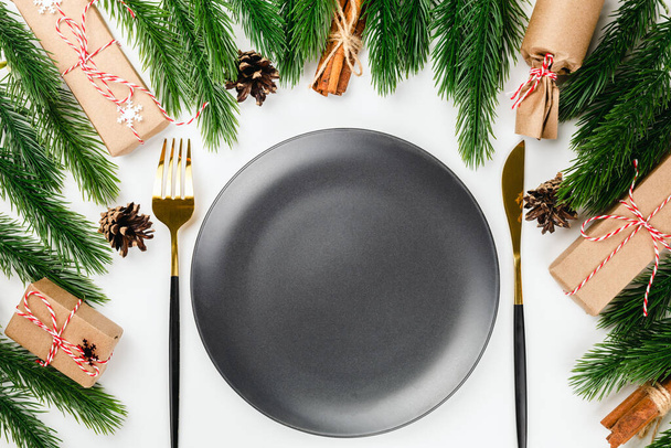 servizio festivo di tavola natalizia con piatto nero, posate in oro e decorazioni fatte di rami di abete, scatole regalo su tavolo bianco - Foto, immagini