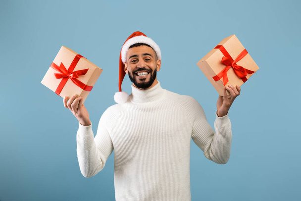 Happy arab guy in santa hat holding christmas or new year gifts and smiling at camera, standing over blue background - Photo, Image