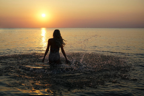 Back view of carefree young woman running into sea splashing water at sunset - Foto, afbeelding