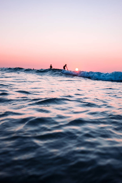 A vertical shot of two people surfing in the ocean at sunset - Φωτογραφία, εικόνα