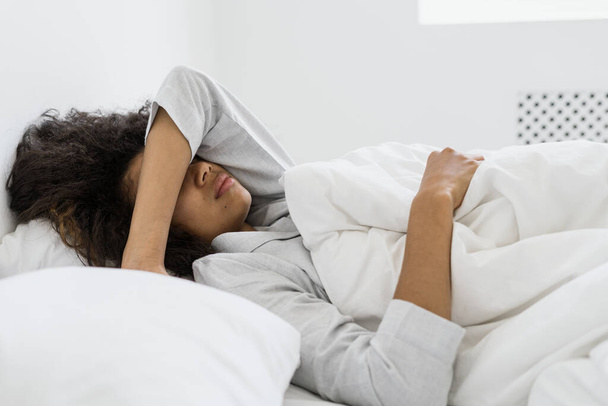 Side view of sad african american woman covered face with hand. Tired dark skinned female lying on bed in pajamas, spending early morning at home, sleeping after stressful night - Φωτογραφία, εικόνα