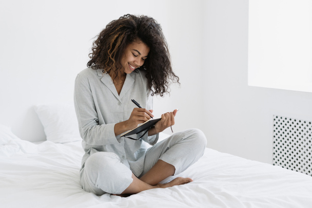 インスピレーション、想像力の概念。若いアフロ系アメリカ人の女性が寝姿でベッドに座り、個人の日記に書いているのを笑っている。幸せな学生作るノートで日記支出朝に自宅 - 写真・画像