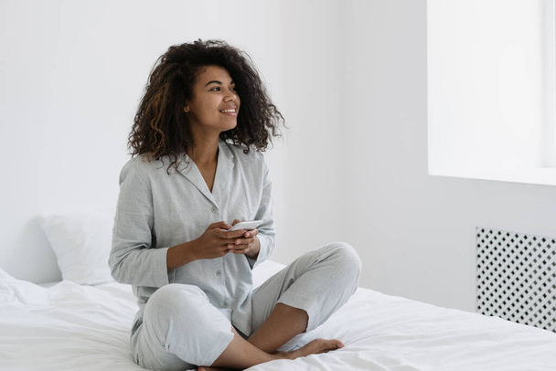 Koncepcja technologii cyfrowej. Szczęśliwa Afroamerykanka korzystająca z nowoczesnego smartfona, uśmiechnięta, szeroka wiadomość tekstowa online. Dziewczyna siedzi na łóżku w piżamie i spędza leniwy weekend rano w domu - Zdjęcie, obraz