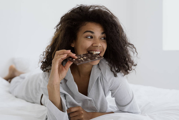 Lezzetli yemek konsepti. Mutlu Afrikalı Amerikalı kadın kahverengi İsviçre çikolatası yiyor, uyku tulumuyla yatağa uzanıyor ve aydınlık bir sabahı evde geçiriyor. Milenyum kızı tatlı tatlının tadını çıkarıyor. - Fotoğraf, Görsel