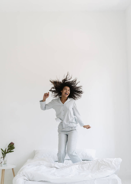 Vertical view of happy young afro american woman jumping up on bed, dance in white bedroom interior. Excited girl wearing in nightwear having fun at home, spend morning, listening music, smiling wide - Foto, Imagem