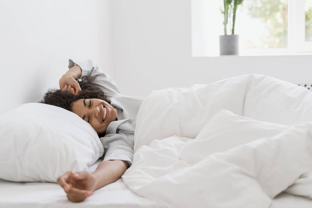Gesunder Schlaf, Erholung und Entspannungskonzept. Glückliche junge Afroamerikanerin, die im weißen Schlafzimmer aufwacht, die Arme ausstreckt und in Schlafanzügen auf dem Bett liegt. Millennial Mädchen verbringen frühen Morgen zu Hause - Foto, Bild
