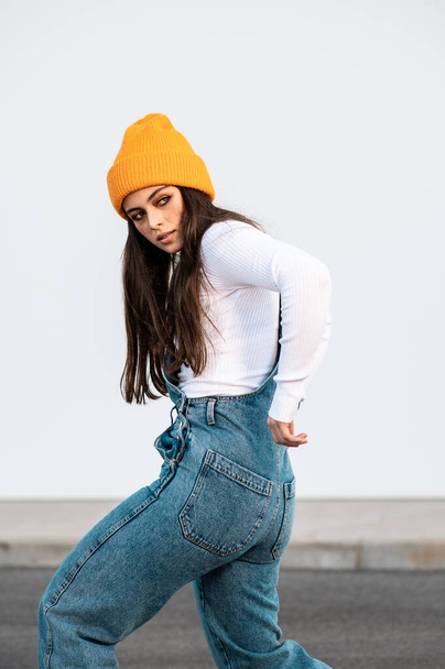 A young Caucasian female wearing denim pants, white shirt and a yellow hat posing against a white wall - Foto, afbeelding