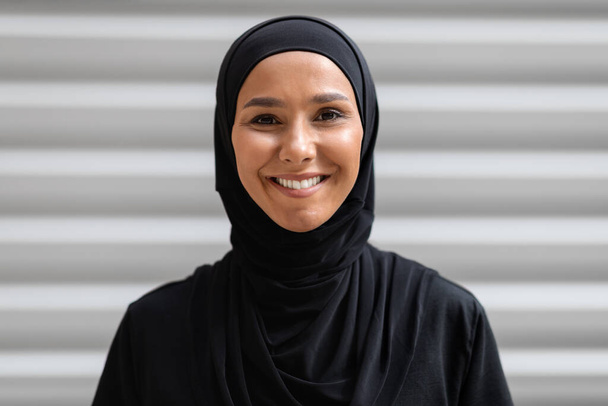 Porträt einer lächelnden jungen arabischen Frau, die einen schwarzen Hijab trägt und im Freien posiert - Foto, Bild