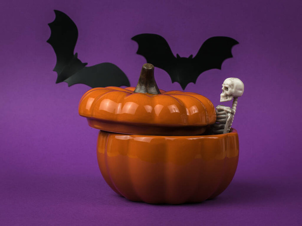 Uno scheletro sporgente da una zucca di ceramica su uno sfondo viola con pipistrelli. Una popolare festa di ottobre. - Foto, immagini