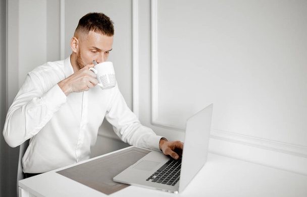 Um homem bebe café trabalhando em um laptop em sua cozinha - Foto, Imagem
