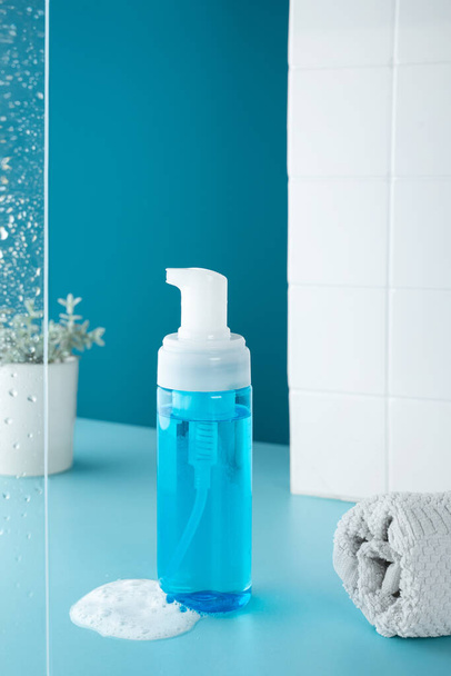 Beauty product mockup. Wellness packaging. Blue bottle, Cleaning cosmetic. Plastic pump bottle for and mousse or cleansing foam. skincare routine. Bath background, glass with drops and a white towel. - Foto, Imagen