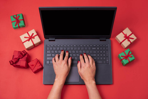 Γυναικεία δακτυλογράφηση χεριών σε laptop με δώρα σε κόκκινο φόντο, εργασία για τις διακοπές, Χριστούγεννα έννοια, κοντινό πλάνο. - Φωτογραφία, εικόνα
