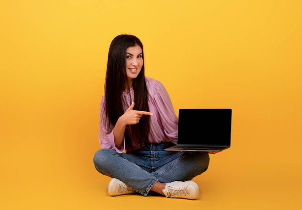 クールなウェブサイトや広告。幸せなアルメニア人の女性がノートパソコンを指差して、黒い画面で、黄色の背景に座って - 写真・画像