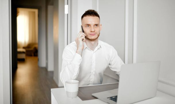 A young man works in the kitchen, talks on the phone, working at a laptop - Photo, Image
