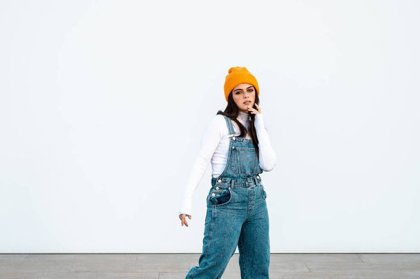 A young Caucasian female wearing denim pants, white shirt and a yellow hat posing against a white wall - Фото, зображення