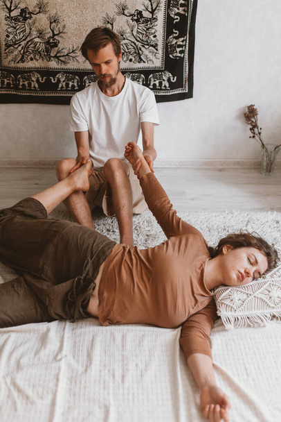 Ein junger bärtiger kaukasischer Masseur und eine Frau machen Thai-Yoga-Massageübungen auf einer Matte zu Hause - Foto, Bild