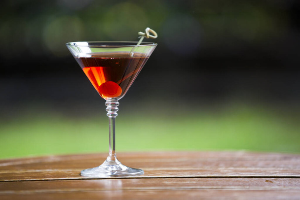 A closeup shot of a glass of red alcohol cocktail with a cherry on a wooden barrel - Фото, изображение