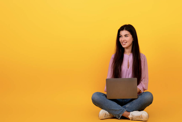 Happy armenian woman using laptop computer on yellow studio background, sitting on floor and looking at copy space - Photo, Image