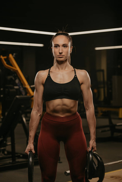 Средний снимок мускулистой женщины, позирующей с двумя резиновыми пластинами в спортзале. Подтянутая девушка в майке и спортивные леггинсы делает тренировку с железными пластинами. - Фото, изображение