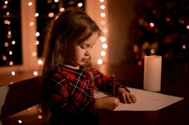 hermosa niña preescolar en la noche en casa escribe una carta para Santa Claus, pide un deseo y regalos. Árbol de Navidad, guirnaldas, luces de Navidad. Esperando vacaciones, calendario de adviento - Foto, imagen