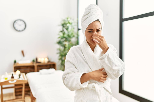 Young brunette woman wearing towel and bathrobe standing at beauty center smelling something stinky and disgusting, intolerable smell, holding breath with fingers on nose. bad smell  - Foto, Imagem