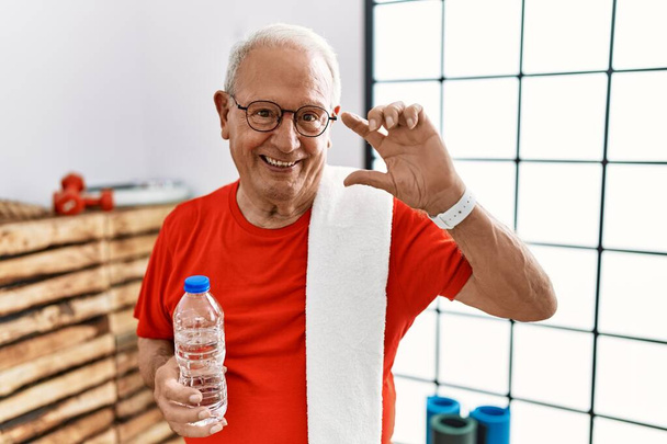 Homme âgé portant des vêtements de sport et une serviette à la salle de gym geste souriant et confiant avec la main faisant signe de petite taille avec les doigts à la recherche et la caméra. concept de mesure.  - Photo, image