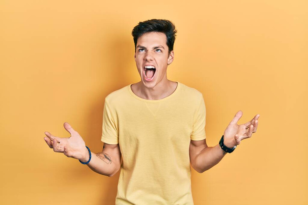 Junger hispanischer Mann in lässigem gelben T-Shirt, verrückt und verrückt schreiend und schreiend mit aggressivem Gesichtsausdruck und erhobenen Armen. Frustrationskonzept.  - Foto, Bild