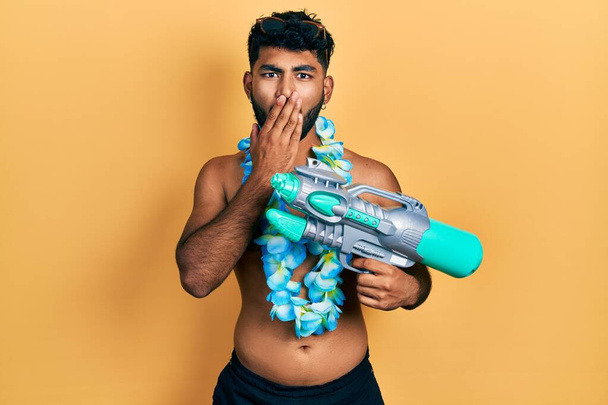 Arab man with beard wearing swimsuit and hawaiian lei holding watergun covering mouth with hand, shocked and afraid for mistake. surprised expression  - Photo, Image