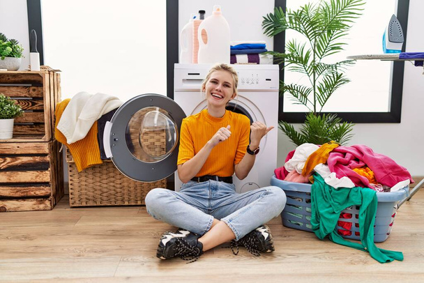 Young blonde woman doing laundry sitting by washing machine pointing to the back behind with hand and thumbs up, smiling confident  - Foto, imagen