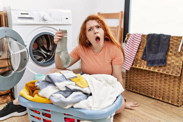 Giovane donna rossa che mette il bucato sporco in lavatrice in faccia shock, guardando scettico e sarcastico, sorpreso a bocca aperta  - Foto, immagini