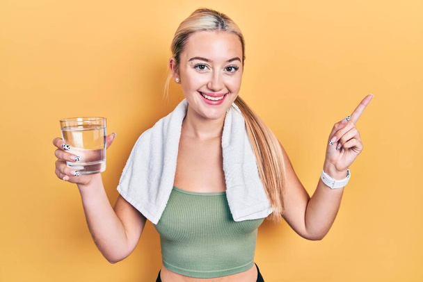 Chica rubia joven con ropa deportiva beber vaso de agua sonriendo feliz señalando con la mano y el dedo a un lado  - Foto, imagen