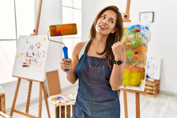 jong latijn kunstenaar vrouw houden verf roller staan op kunst studio duim wijzen naar de zijkant glimlachen blij met open mond  - Foto, afbeelding