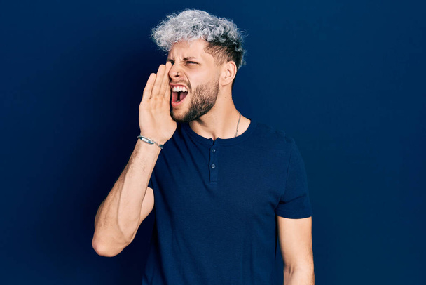 Молодий іспаномовний чоловік з сучасним пофарбованим волоссям в повсякденну синю сорочку, що кричить і кричить голосно на бік з рукою на рот. концепція спілкування
.  - Фото, зображення
